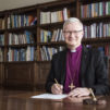 harmaahiuksinen-mies-jolla-liila-piispanpaita-päällään-istuu-kirjoituspöydän ääressä ja hymyilee