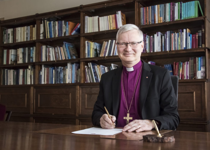 harmaahiuksinen-mies-jolla-liila-piispanpaita-päällään-istuu-kirjoituspöydän ääressä ja hymyilee
