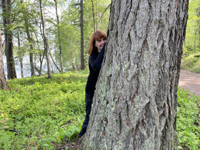 vihreässä-luonnossa-kuvan-keskellä-iso-puun-runkoä-jonka-takaa-kurkistaa-tummahiuksinen-keski-ikäinen-nainen