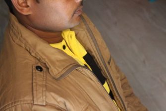 Kuvassa on Shekhar sivusta päin kuvattuna. Kuva on rajattu niin, että hänestä näkyy vain korvan puolivälistä alaspäin kasvot. Hänellä on mustat hennot viikset ja beige takki, jonka alla kelta-mustaraidallinen paita.
