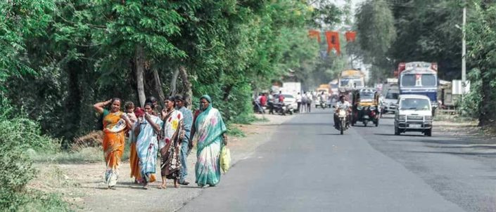 Puiden lomassa kulkevan autotien vierellä kävelee useita intialaisia naisia. Tiellä ajetaan monenlaisilla menopeleillä.
