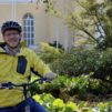 keski-ikäinen-mies-keltainen-takki-päällä-ja-pyöräilykypärä-päässä-pyörän-luona-taustalla-keltainen-rakennus-ja-vihreää-luontoa