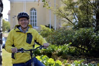 keski-ikäinen-mies-keltainen-takki-päällä-ja-pyöräilykypärä-päässä-pyörän-luona-taustalla-keltainen-rakennus-ja-vihreää-luontoa