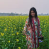 Nuori tummahiuksinen nainen seisoo värikkäässä asussa keskellä peltoa joka on täynnä keltaisia kukkia.