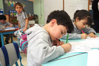 lapsi istuu pulpetin ääressä koululuokassa ja kirjoittaa paperiin