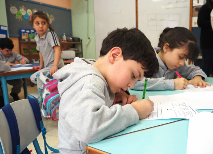 lapsi istuu pulpetin ääressä koululuokassa ja kirjoittaa paperiin