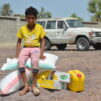 Jemeniläinen poika istuu ruoka-apu kassien ja kanistereiden päällä keltaisessa paidassa. Takana näkyy valkoinen auto.