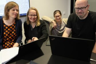 Neljä ihmistä istuvat pöydän ääressä ja katsovat kannettavan tietokoneen näyttöä