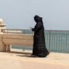 Mustiin pukeutunut nainen kävellee rantakadulla veden äärellä älypuhelin kädessä.