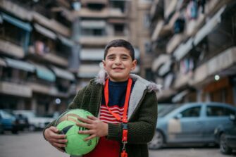 Poika hymyilee vihreä pallo käsissään. Hän on rakennusten ja autojen keskellä.