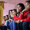 Tummatukkaiset alakouluikäiset tytöt istuvat vinosti vierekkäin ja rivin päässä näkyy heidän naispuolinen pyhäkouluopettajansa. Huoneen seinät ovat vaaleanpunaiset ja verhot keltaiset.