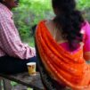 Kauluspaitainen mies ja oranssiin sariin pukeutunut pitkähiuksinen nainen istuvat selkäpäin kameraan penkillä.