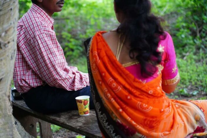 Kauluspaitainen mies ja oranssiin sariin pukeutunut pitkähiuksinen nainen istuvat selkäpäin kameraan penkillä.
