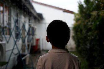 Mustatukkainen pikkupoika valkoisessa paidassa selkäpäin talon vieressä.