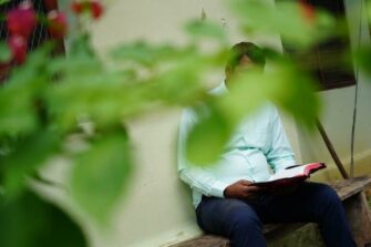 Intialainen Vijay lukee Raamattua vaaleansinisessä paidassa ulkona penkillä seinän vieressä. Kuvassa etualalla oleva vihreälehtinen oksa peittää hänen kasvojaan.