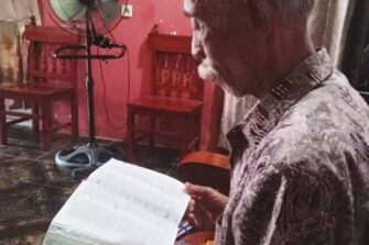 Harmaantunut mies lukee raamattua istualtaan sivuttain kameraan nähden kodissaan