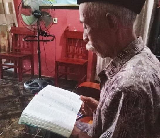 Harmaantunut mies lukee raamattua istualtaan sivuttain kameraan nähden kodissaan