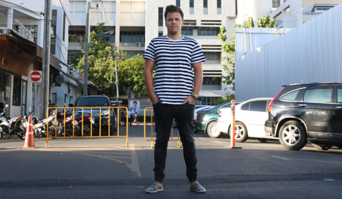 Mies seisoo parkkipaikalla mustavalkoraidallisessa t-paidassa ja mustissa farkuissa.