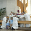 Nelihenkinen perhe on polvistunut altarinne. Heillä on päällään valkean albat. Alttarikaiteen takana on kahdeksan henkilöä, jotka ovat ojentaneet kätensä polvistuneita kohti. Keskellä seisoo piispa sauvoineen.