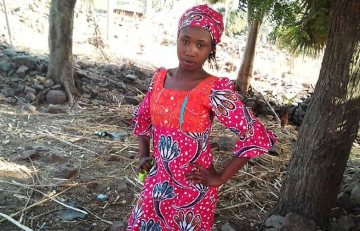 Teini-ikäinen nigerialainen tyttö, Leah Sharibu, kädet lanteillaan. Hänellä on punaisenkirjava mekko ja huivi.