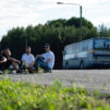 neljä miestä istuu skeittilautojen päällä maantiellä, takana näkyy huoltobussi