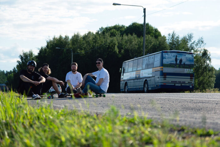 neljä miestä istuu skeittilautojen päällä maantiellä, takana näkyy huoltobussi