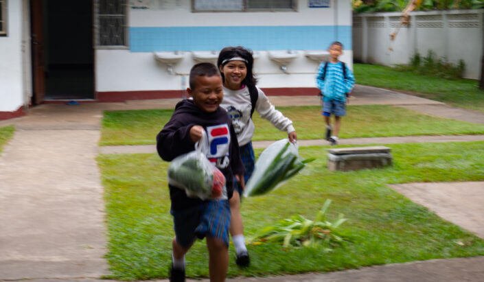 Kaksi thaimaalaista lasta juoksee muovipussit kädessä joissa on vihanneksia.