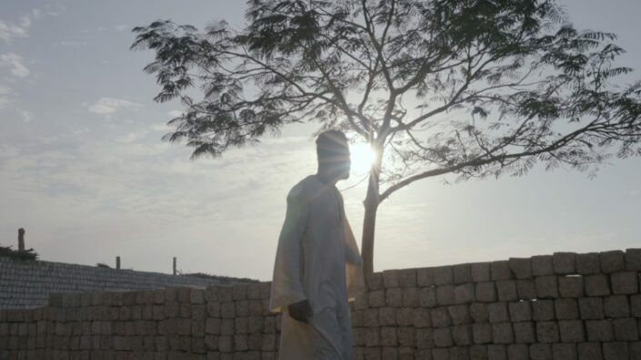 Mies seisoo valkeissa vaatteissa puun ja auringonvalon edessä.