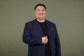Mongolialainen mustaan pukuun pukeutunut mies hymyilee kameraan