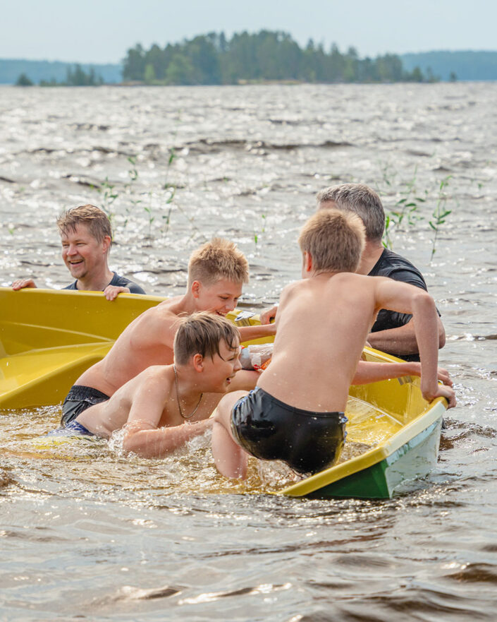 kaksi miestä seisoo rantavedessä ja keinuttaa venettä, jossa on pieniä poikia