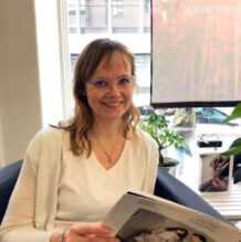 Nainen, Johanna Lukkarila, istuu työpöydän ääressä lehti käsissään.