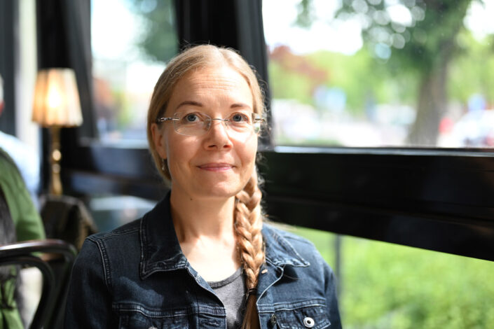 Ulla Barendsen-Riikonen istuu pöydässä ikkunan äärellä. Hänen hiuksensa ovat letillä, ja hänellä on silmälasit.