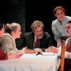 Pöydän ääressä istuu näytelmän hahmoja syventyneenä tiiviiseen keskusteluun. Kolme hahmoista on nuorehkoja naisia, kaksi muuta vanhempia herroja.