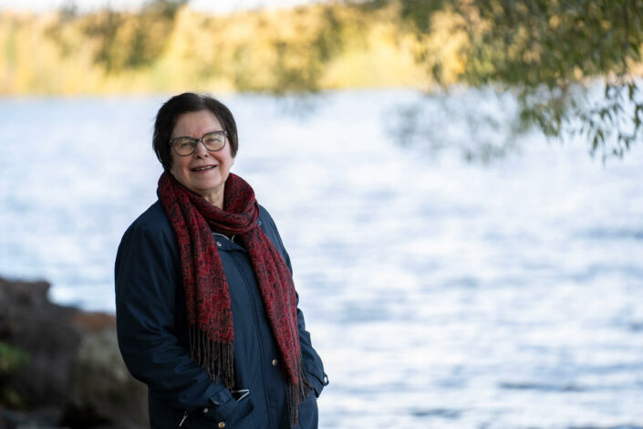 Marketta Takku seisoo vesistön äärellä ja hymyilee.