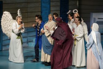 Teatterin näyttämöllä ryhmä näyttelijöitä rooliasuissaan, Ami Aspelund valkoisessa, pitkässä vaatteessa, selässään enkelin siivet.