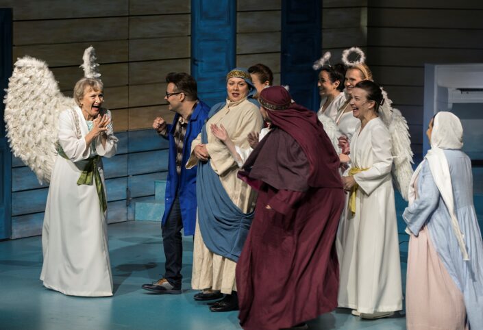 Teatterin näyttämöllä ryhmä näyttelijöitä rooliasuissaan, Ami Aspelund valkoisessa, pitkässä vaatteessa, selässään enkelin siivet.