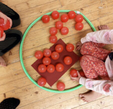 Kuvassa on kaksi lasta ylhäältä kuvattuna. Lasten kasvot eivät näy. Lasten välissä on hulahula-rengas, jonka sisäpuolelle on koottu punaisia muovipalloja.