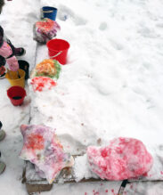 Kuvassa on lumeen peittynyt hiekkalaatikko, jonka reunalla on ämpäreitä ja lumikökkäreitä, joita on maalattu ämpäreissä olevilla väreillä.