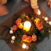 Kuvan keskellä on suuri kynttilä, ja sen ympärillä oransseja ruusuja, havuja, pienempiä kynttilöitä sekä lasisia timantteja.