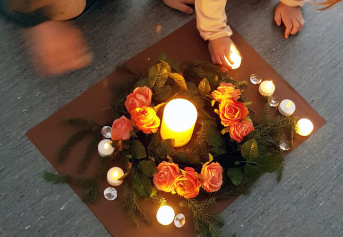 Kuvan keskellä on suuri kynttilä, ja sen ympärillä oransseja ruusuja, havuja, pienempiä kynttilöitä sekä lasisia timantteja.