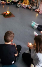 Kuvassa on lapsia ringissä lattialla istumassa ledikynttilät käsissään. Lasten kasvot eivät näy. Ringin keskellä on asetelma, jossa on kynttilä, ruusuja ja havoja ruskealla pohjalla.