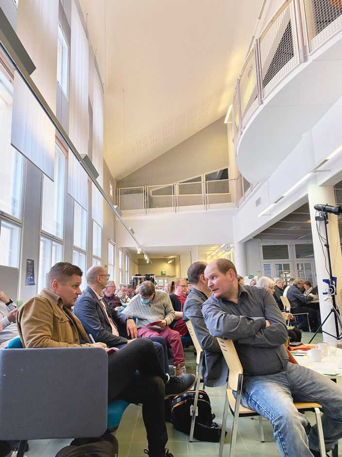 Kuvassa on vaalea, korkea tila, jossa istuu kymmeniä ihmisiä. etualalla on kaksi miestä, jotka keskustelevat keskenään.