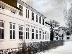 Kuvassa on sivulta kuvattu vanha kartano, jonka takana näkyy toinen rakennus. Molemmat talot ovat vaaleita. Kuva on mustavalkoinen. Maassa on lunta.