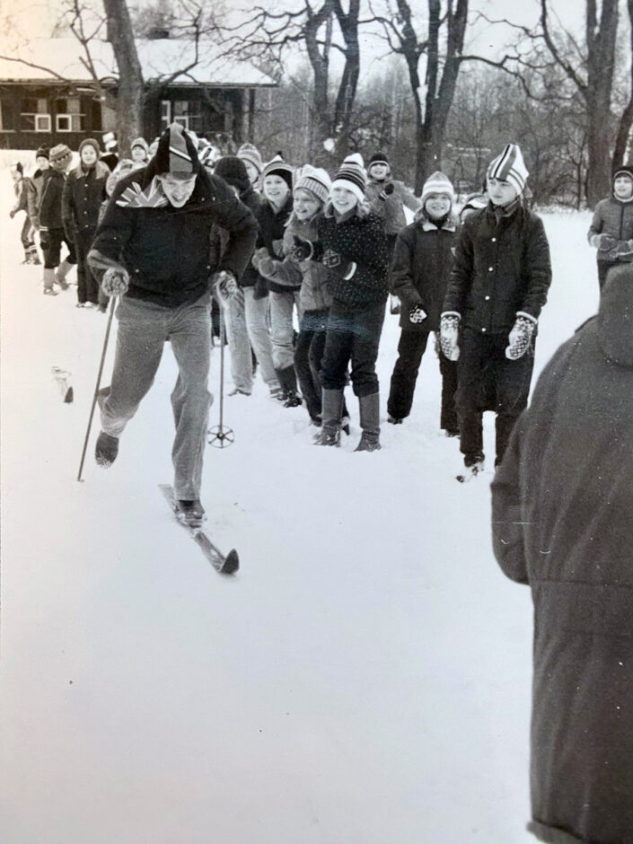Kuvassa on joukko lapsia ja nuoria. Yksi heistä hiihtää innokkaasti kohti kuvaajaa. Kuva on mustavalkoinen.