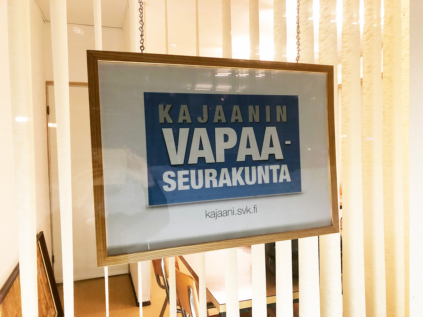 Kuvassa näkyy ikkuna ulkoapäin kuvattuna. Ikkunassa on säleverho ja lisäksi kyltti, jossa on Kajaanin vapaaseurakunnan uusi logo. Se koostuu tekstistä, joka on sinisellä pohjalla valkoisessa kehyksessä.
