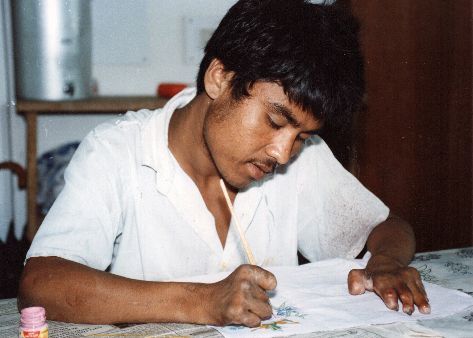 Kuvassa on nuori nepalilaismies, joka piirtää tai kirjoittaa valkoisessa paidassa. Miehellä on ruskea iho ja tummat hiukset ja viikset. Hänen sormensa näyttävät surkastuneilta. Ilme on keskittynyt.