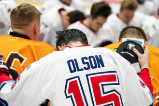 Kuvassa on polvistuneita, rukoilevia jääkiekkoilijoita valkoisissa ja keltaisissa asuissa. Etualalla olevan pelaajan selässä lukee "Olson". Hänen pelinumeronsa on 15.