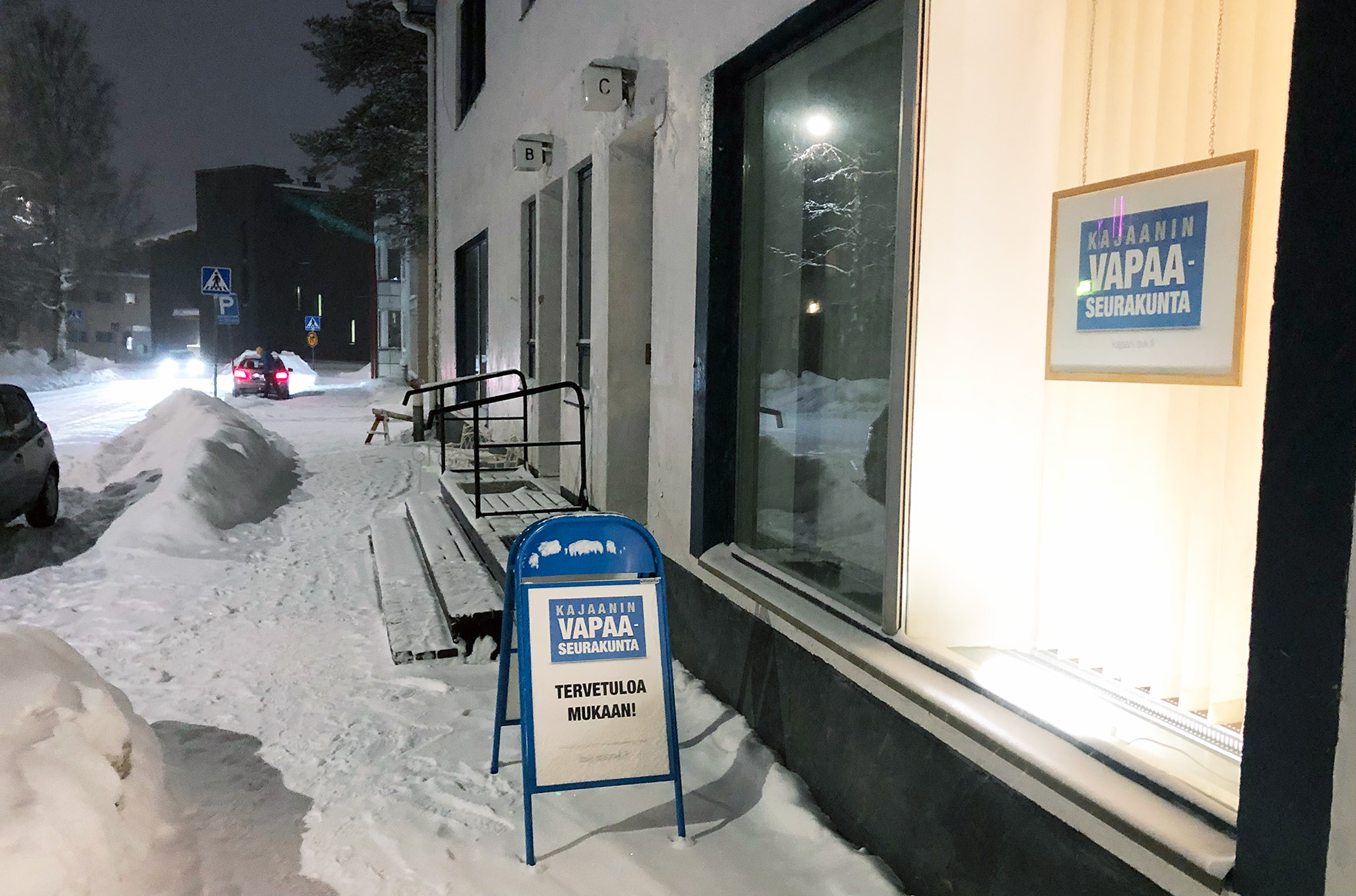 Kuvassa on lumen peittämä katu ja talo, jonka näyteikkunasta kajastaa valo. Näyteikkunassa ja myös kadulla on kyltti, jossa lukee Kajaanin vapaaseurakunta.