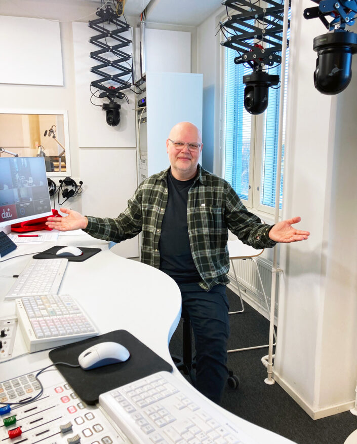 Kuvassa on keski-ikäinen, kalju, vaaleapartainen mies vaaleissa silmälaseissa. Vihreäsävyinen flanelliruutupaita on auki, alla on musta t-paita, jalassa mustat housut. Mies istuu pinnoiltaan valkoisessa studiossa. Kuvan etualalla ja vasemmassa reunassa on pöytää, jolla on studioon liittyvää tekniikkaa. Katosta roikkuu ilmeisesti mustia kameroita. Mies on levittänyt kädet sivuilleen ja hymyilee kainosti.