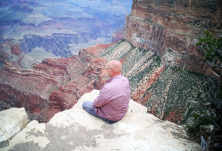 Kuvassa näkyy punertavasävyistä Grand Canyonin maisemaa. Etualalla on vaalea kieleke, jolla istuu marjapuuronvärisessä paidassaan ja vaaleansinisissä housuissaan Mikael Juntunen. Hän eon selin kameraan ja katsoo alhaalla avautuvaan maisemaan.